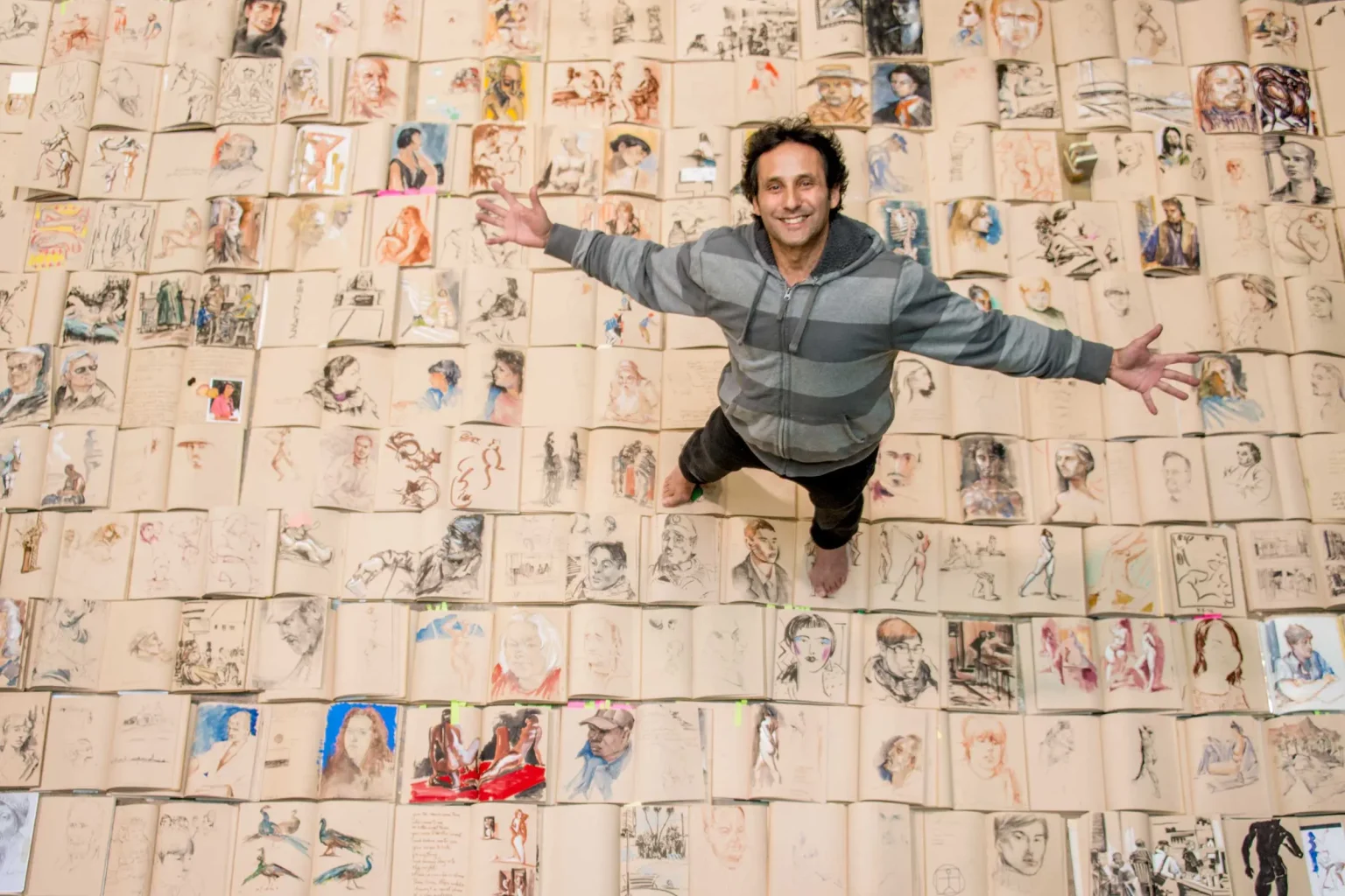 Artist with arms spread wide, standing on a floor covered with open sketchbooks